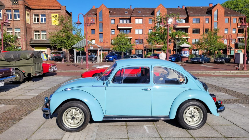 Ein hellblauer Oldtimer-VW-Käfer steht auf einer gepflasterten Fläche, im Hintergrund sind rote Backsteingebäude und andere Autos zu sehen.